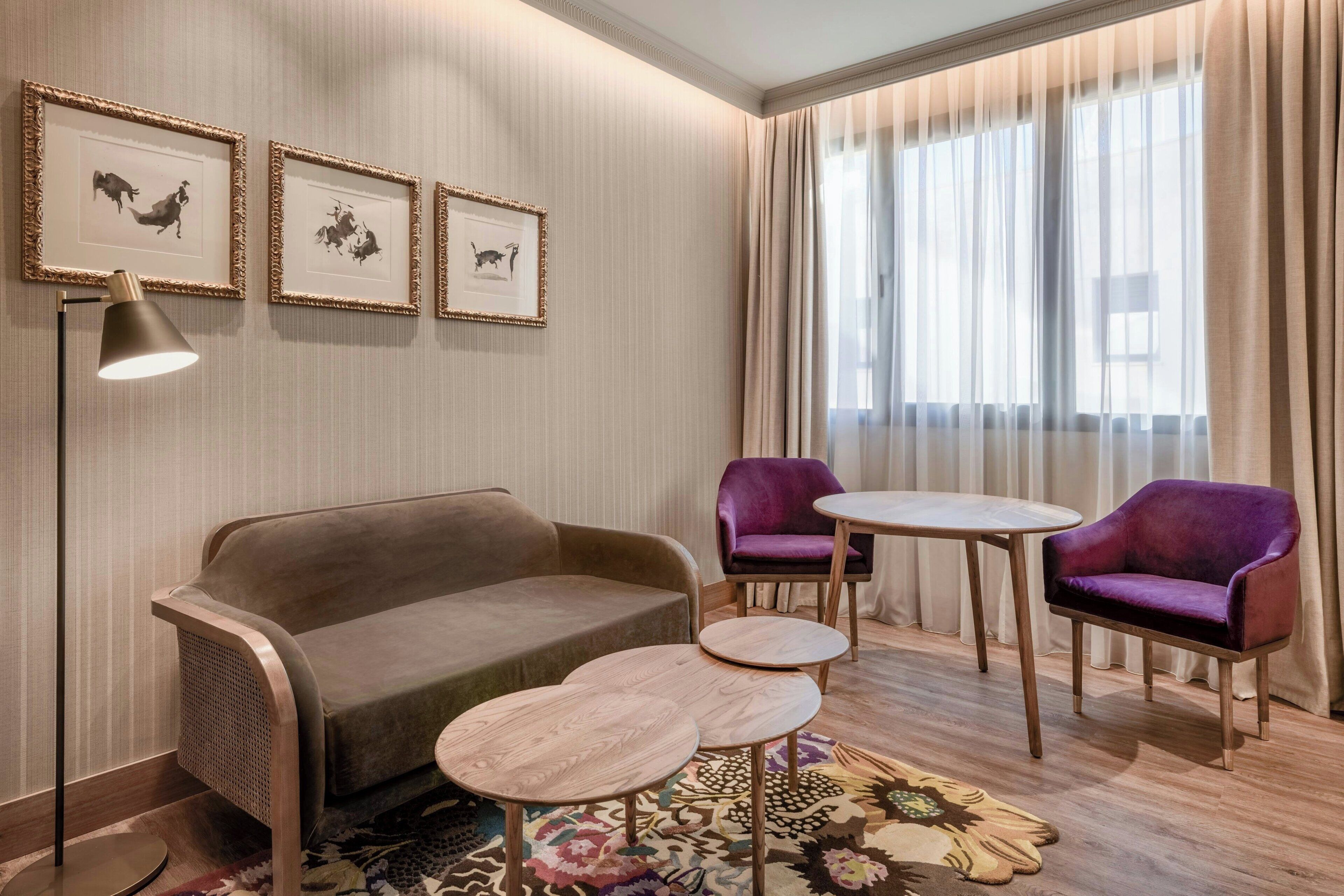A modern hotel room with a sofa, two purple armchairs, a small round table, and framed artwork on the wall.