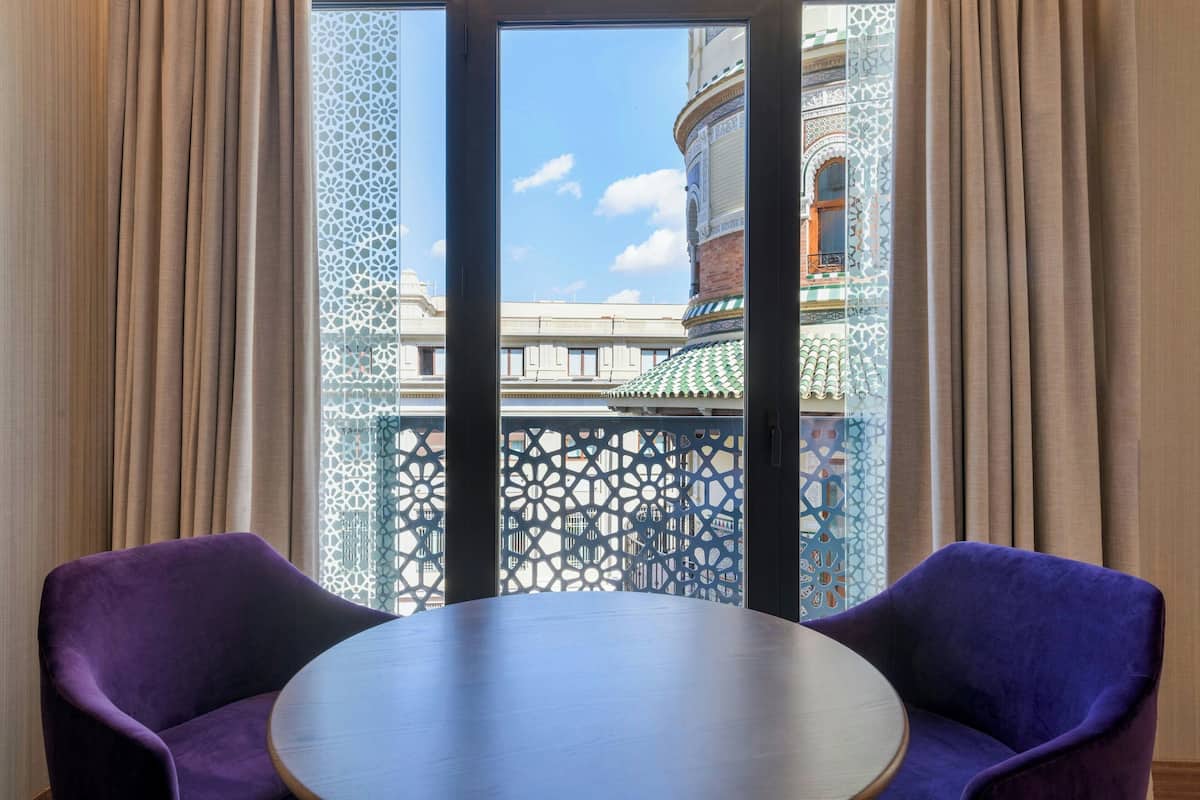 Una habitación con una mesa redonda y dos sillas moradas, un ventanal con un diseño y vistas a un edificio con tejado verde.