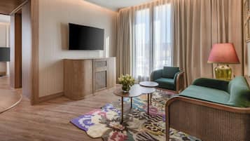 A modern hotel room with a flat-screen TV, a green sofa, a small table with a vase of flowers, and a patterned rug.