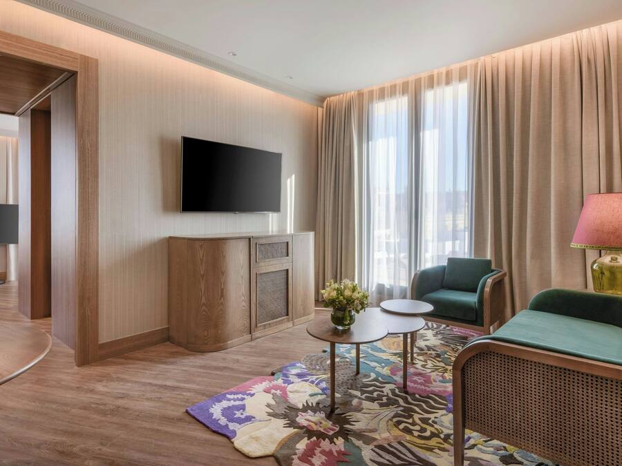 A modern hotel room with a flat-screen TV, a green sofa, a small table with a vase of flowers, and a patterned rug.