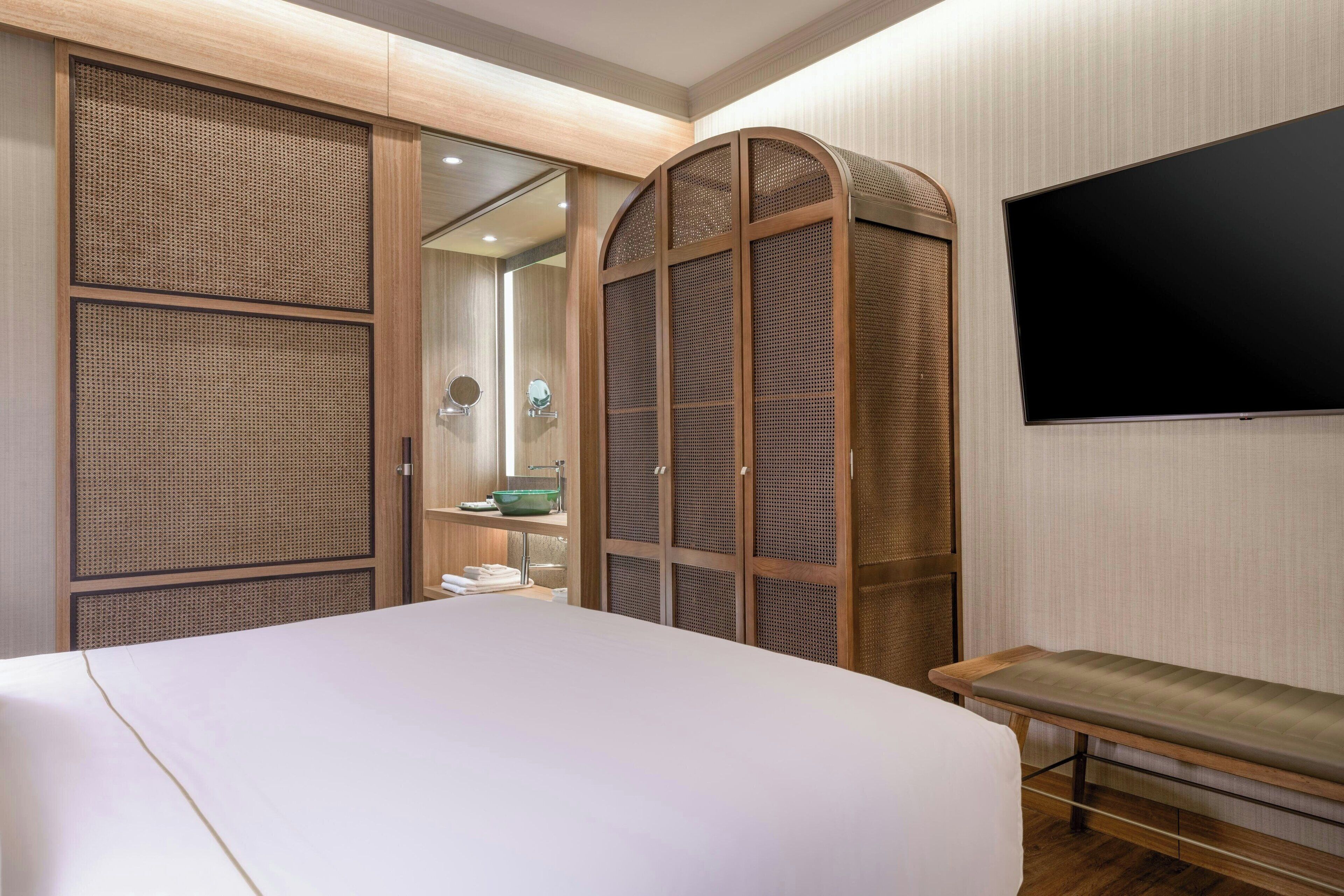 A hotel room with a large bed, a bench, a TV, and a wardrobe with a mirror and a green bowl.