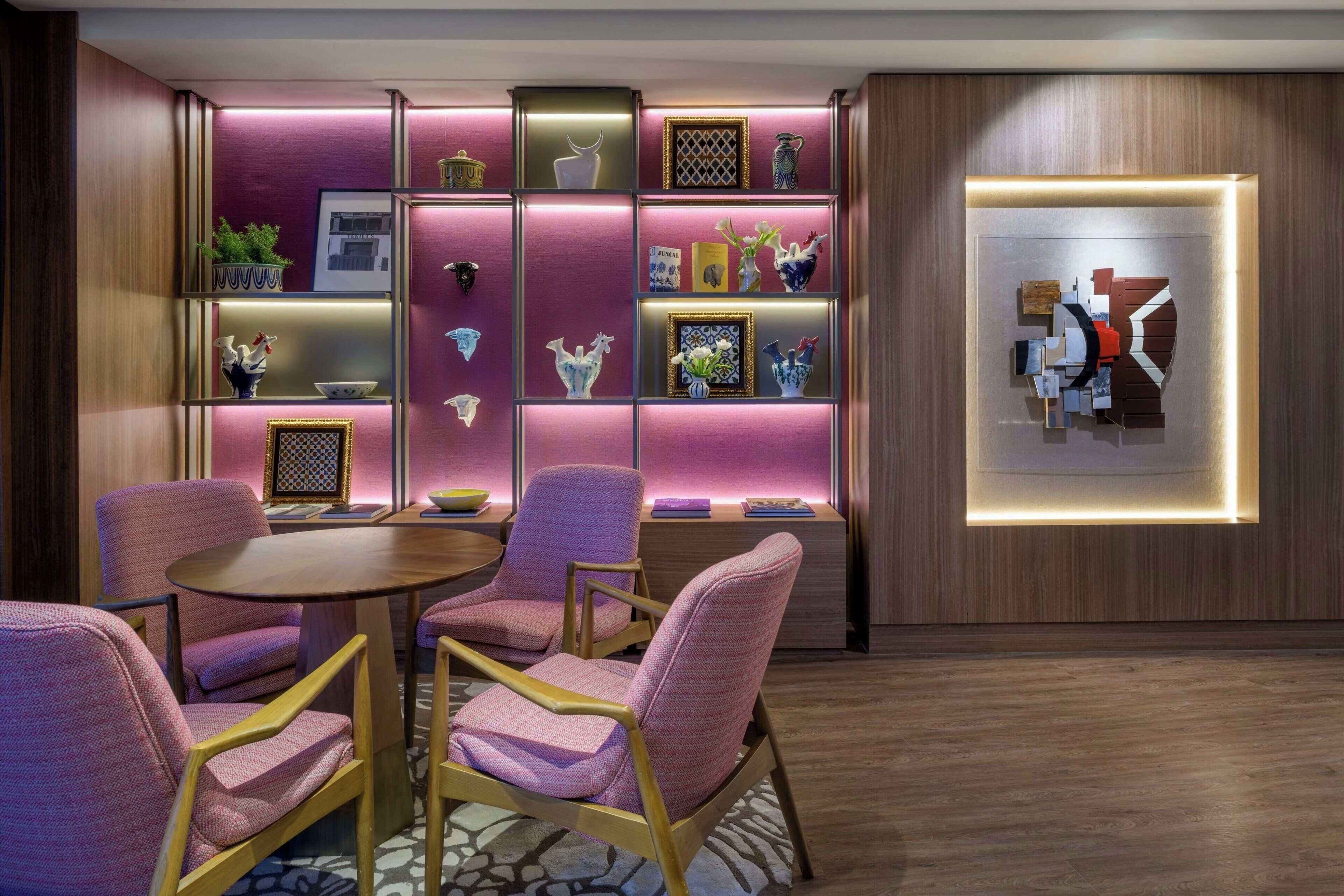A modern room with a wooden floor, a round table, and four pink chairs. There is a wall with shelves holding various decorative items and a framed abstract artwork.