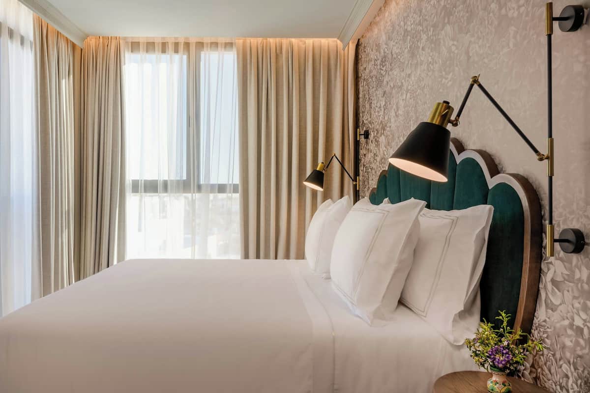 Una habitación de hotel con una cama grande, cortinas color beige, un cabecero tapizado en verde, una lámpara de escritorio negra y un florero.