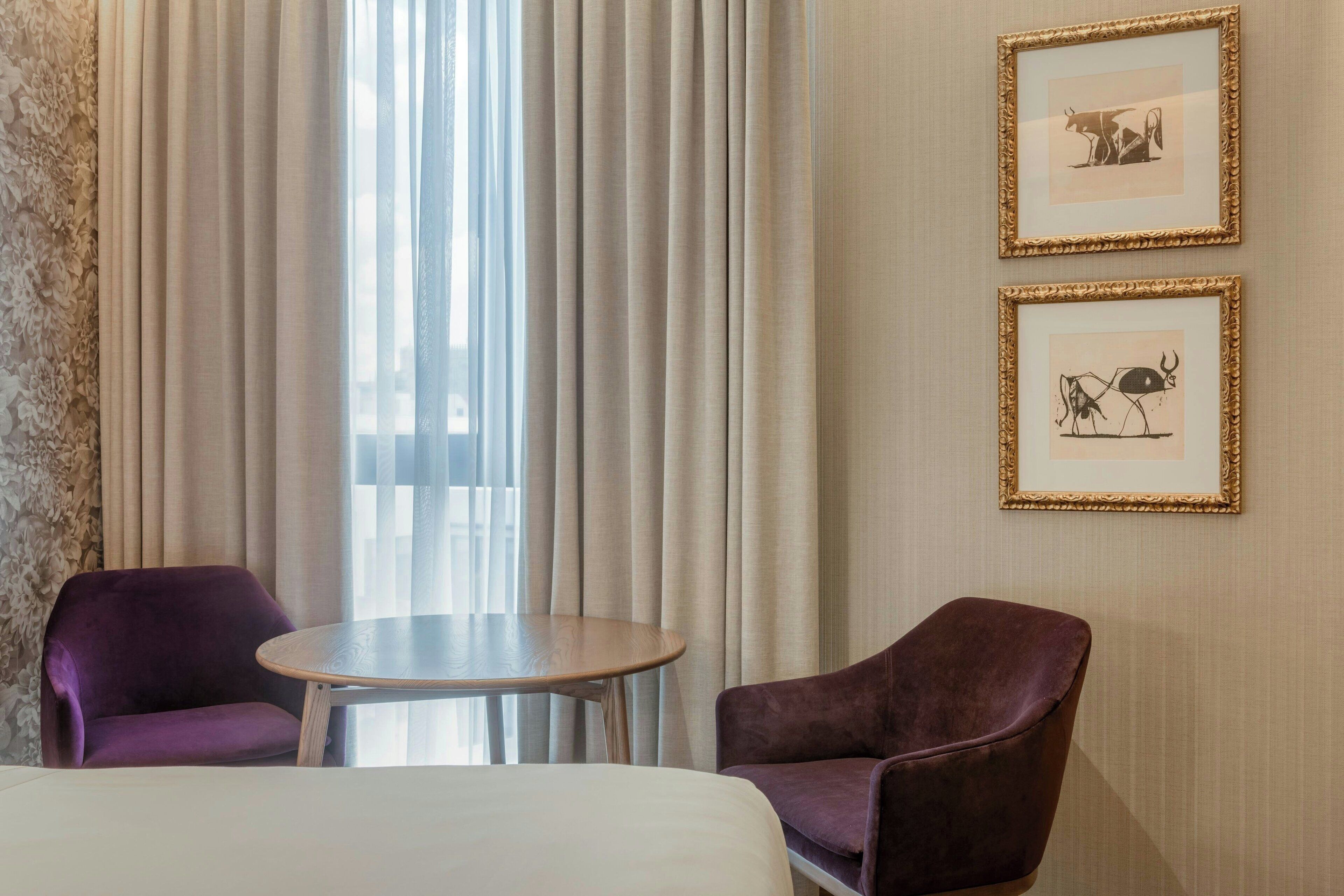 A hotel room with a bed, two purple armchairs, a small round table, and two framed pictures on the wall.
