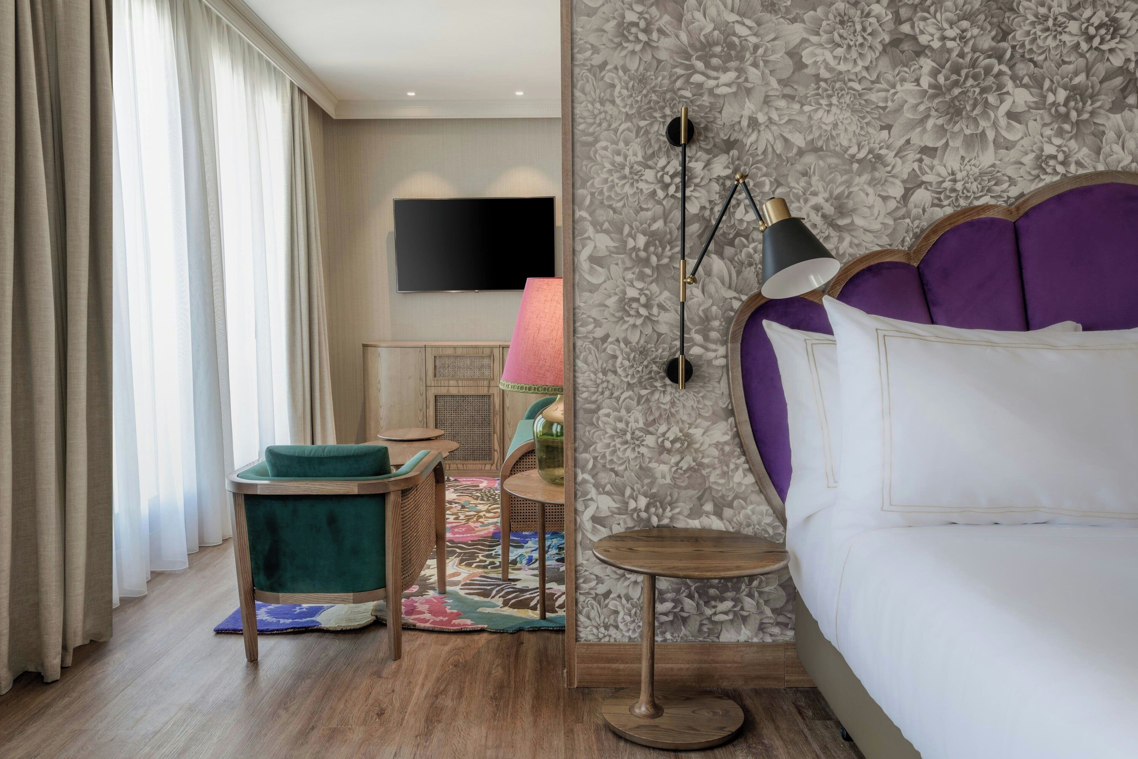 A hotel room with a bed, a TV mounted on the wall, a floor lamp, and a small table with a chair.