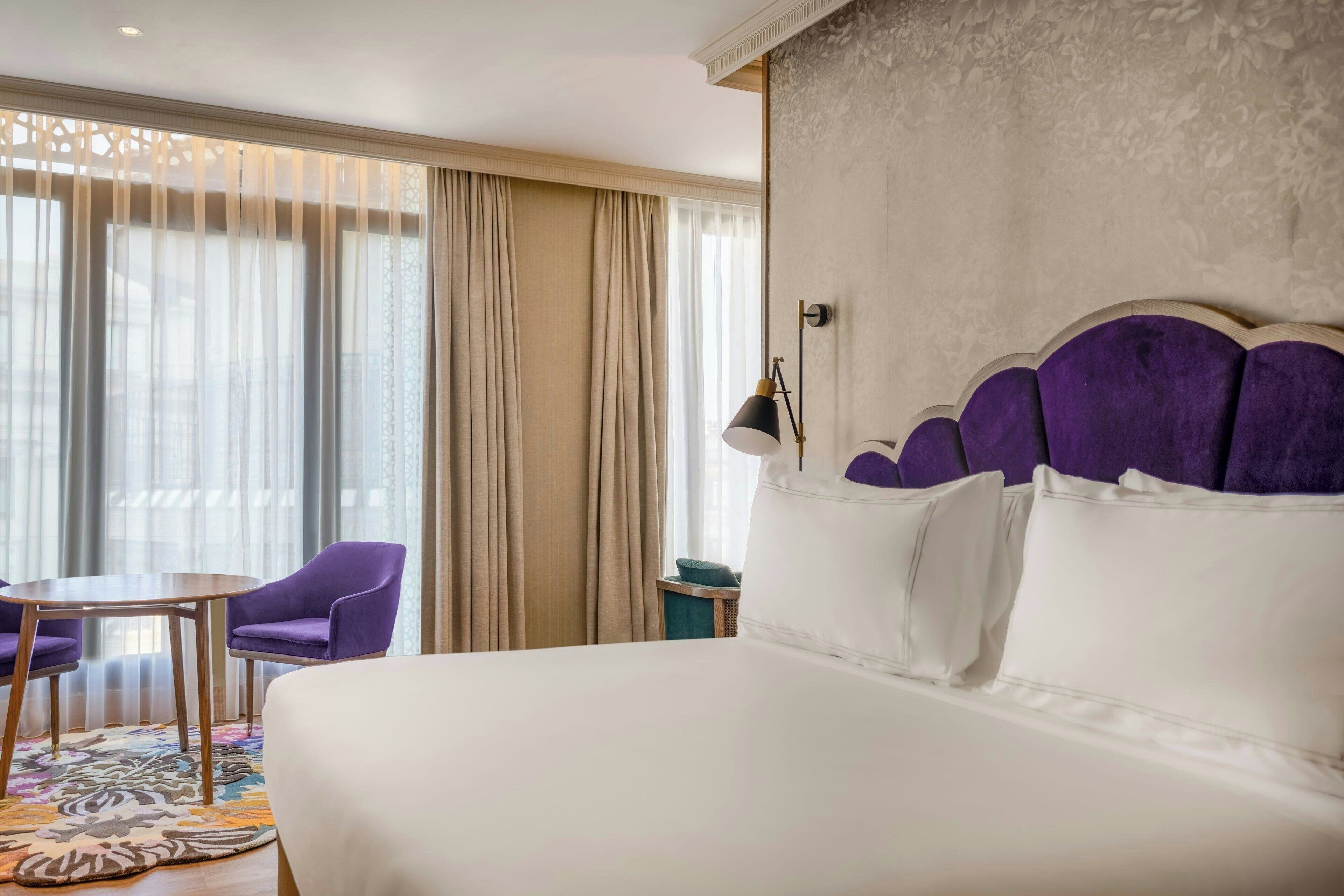 A hotel room with a large bed, a small table, purple chairs, and a view of the city through sheer curtains.