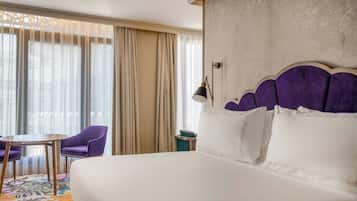 A hotel room with a large bed, a small table, purple chairs, and a view of the city through sheer curtains.