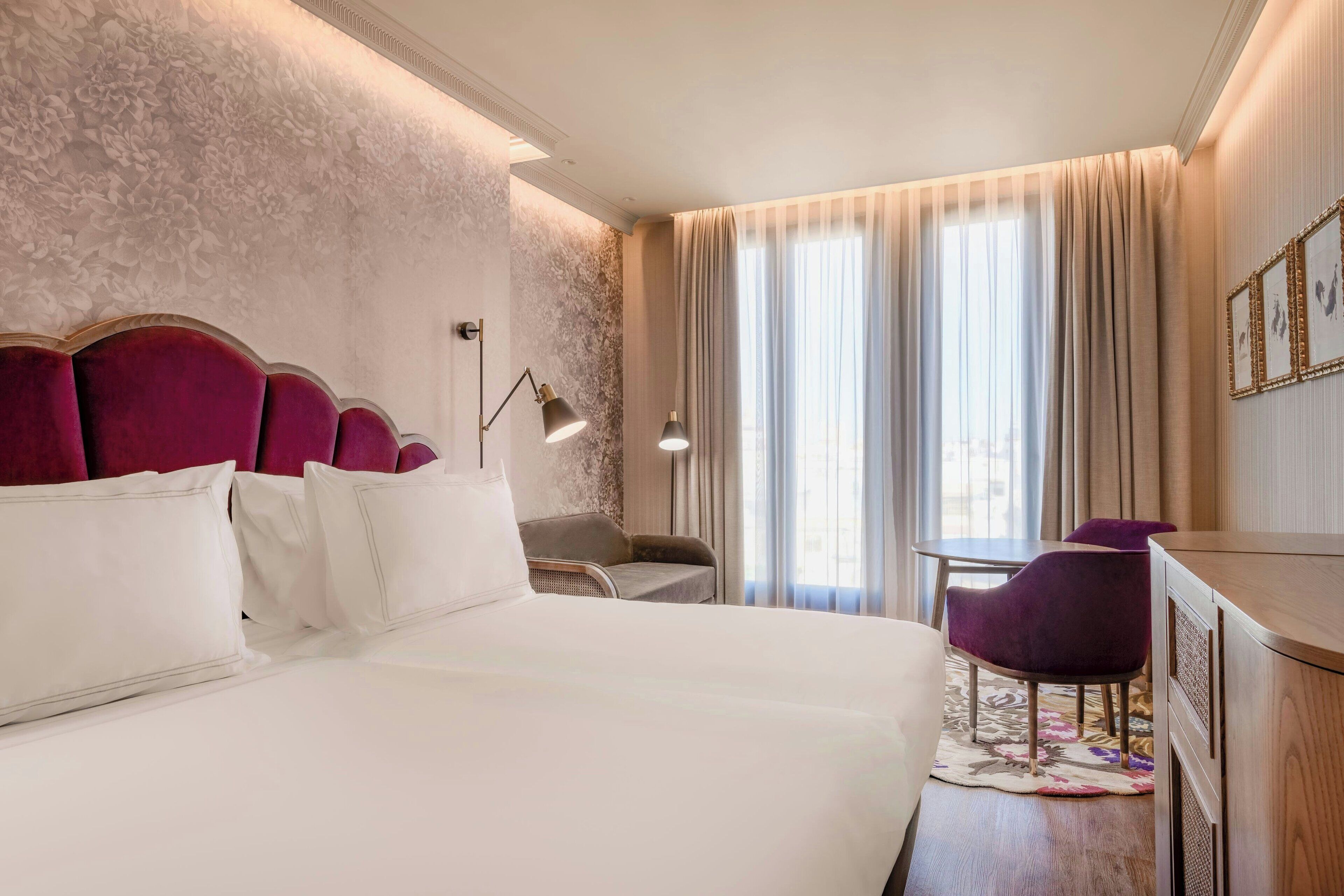 A hotel room with a large bed, a desk, two chairs, and a window with curtains.