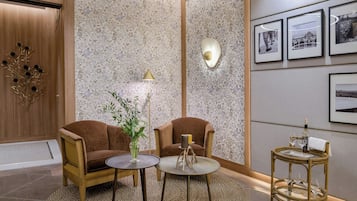 Un hall d’entrée moderne avec deux fauteuils marron, une table ronde ornée d’un vase, un lampadaire et une petite table ronde sur laquelle sont posés des verres et une bouteille.