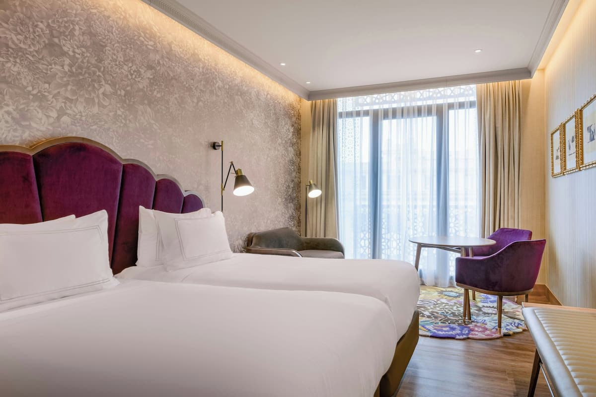 Habitación de hotel con dos camas, una mesita pequeña, sillas moradas y un ventanal grande con cortinas.