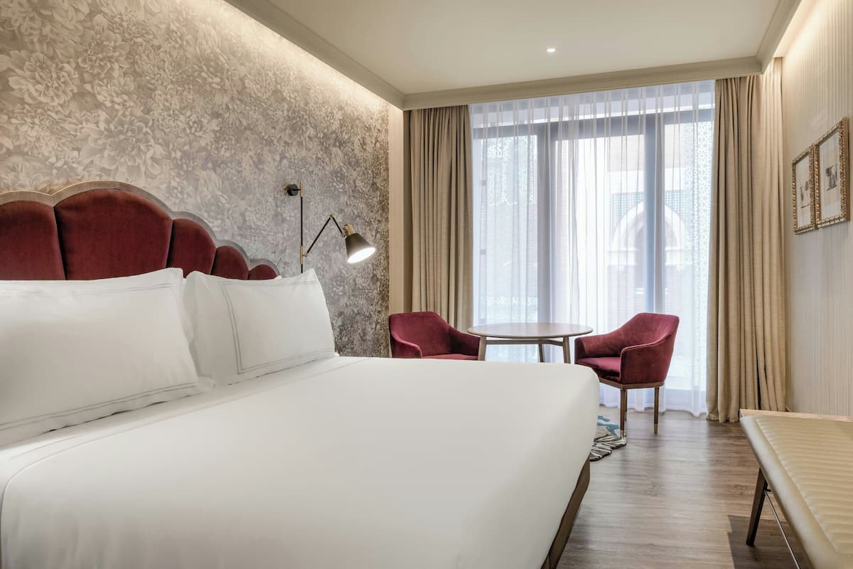 Habitación de hotel con una cama grande, un sillón rojo, una mesita redonda y vistas de la ciudad a través de cortinas traslúcidas.