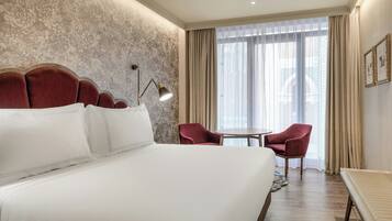 Habitación de hotel con una cama grande, un sillón rojo, una mesita redonda y vistas de la ciudad a través de cortinas traslúcidas.