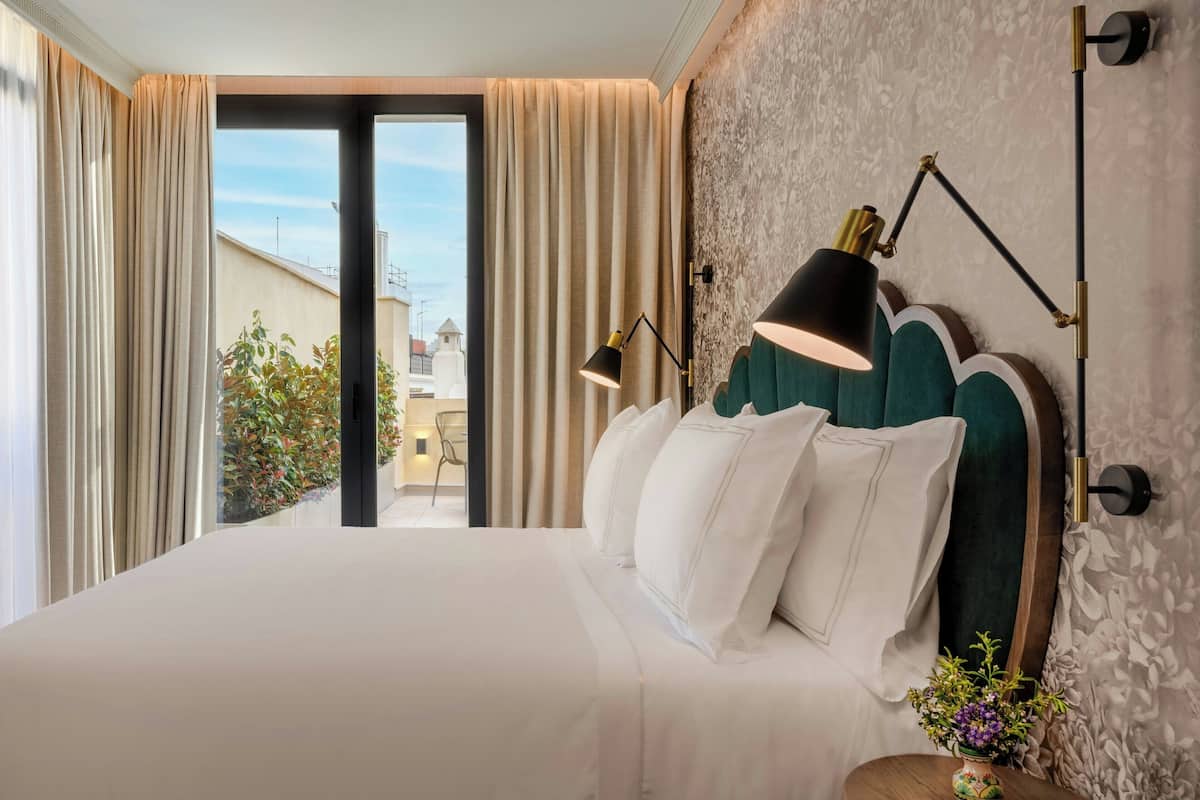Habitación de hotel con una cama grande, dos lámparas de noche, vistas al exterior y una mesita con un jarrón de flores.