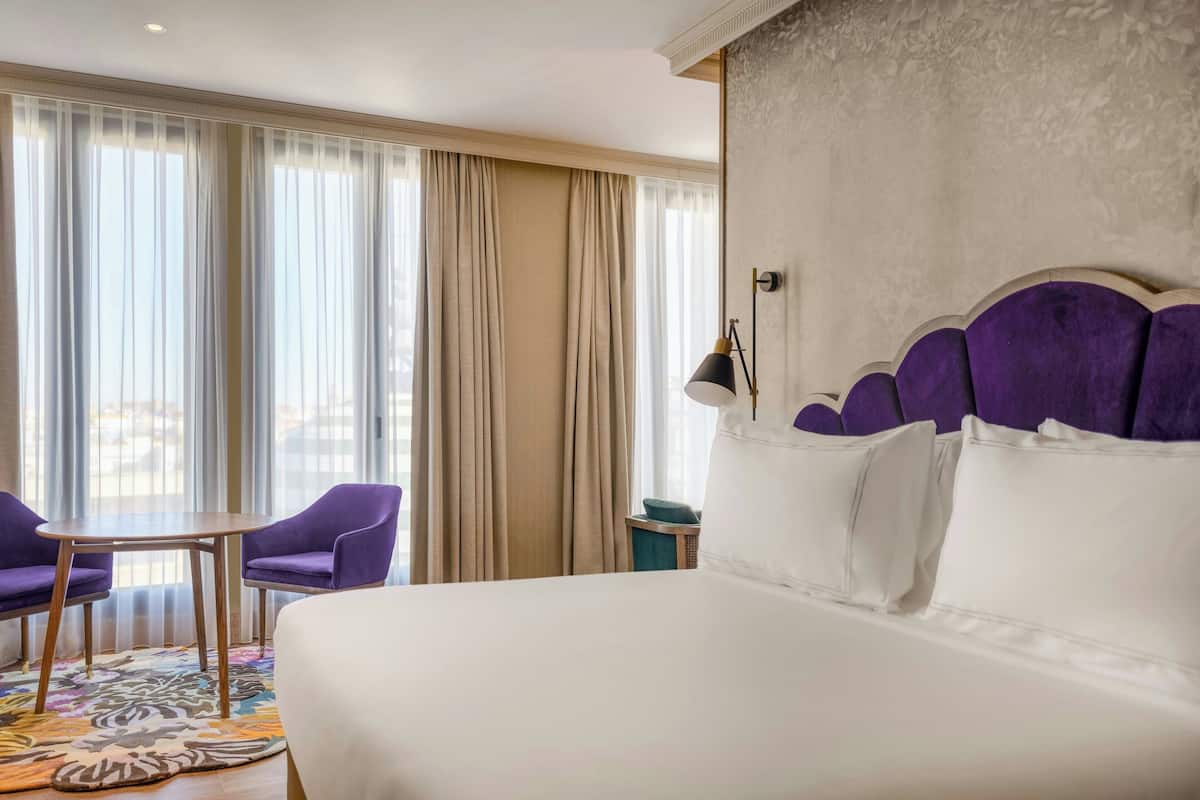 Una habitación de hotel con una cama grande, dos sillas moradas, una mesita redonda y vistas a través de cortinas translúcidas.