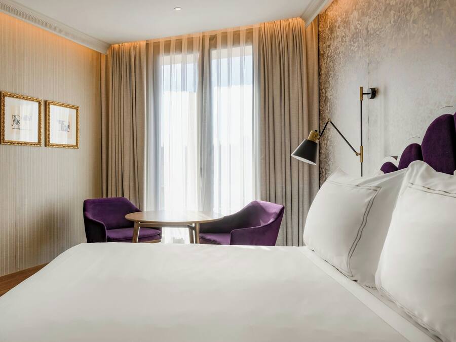 A hotel room with a large bed, two purple chairs, a small table, and a large window with sheer curtains.