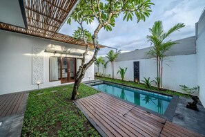 A modern pool area with a wooden deck, a small tree, and a white wall with decorative elements. - The Dewi Villa Canggu (Tumbak Bayuh)