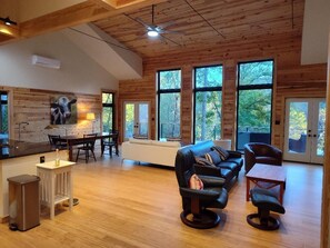 Amplia sala de estar con techo de madera, ventanas grandes con vistas al exterior, un sofá de cuero negro y una zona de comedor con mesa y sillas.