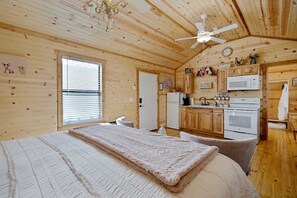 Una habitación acogedora estilo cabaña con cama, ventilador de techo, paredes de madera y kitchenette.