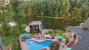 A swimming pool with a deck, lounge chairs, and umbrellas, surrounded by a wooded area and a lake. - The Oasis at Lake Gaston - Heated Pool, Hot Tub, Pontoon Boat Rental Included! (Littleton)
