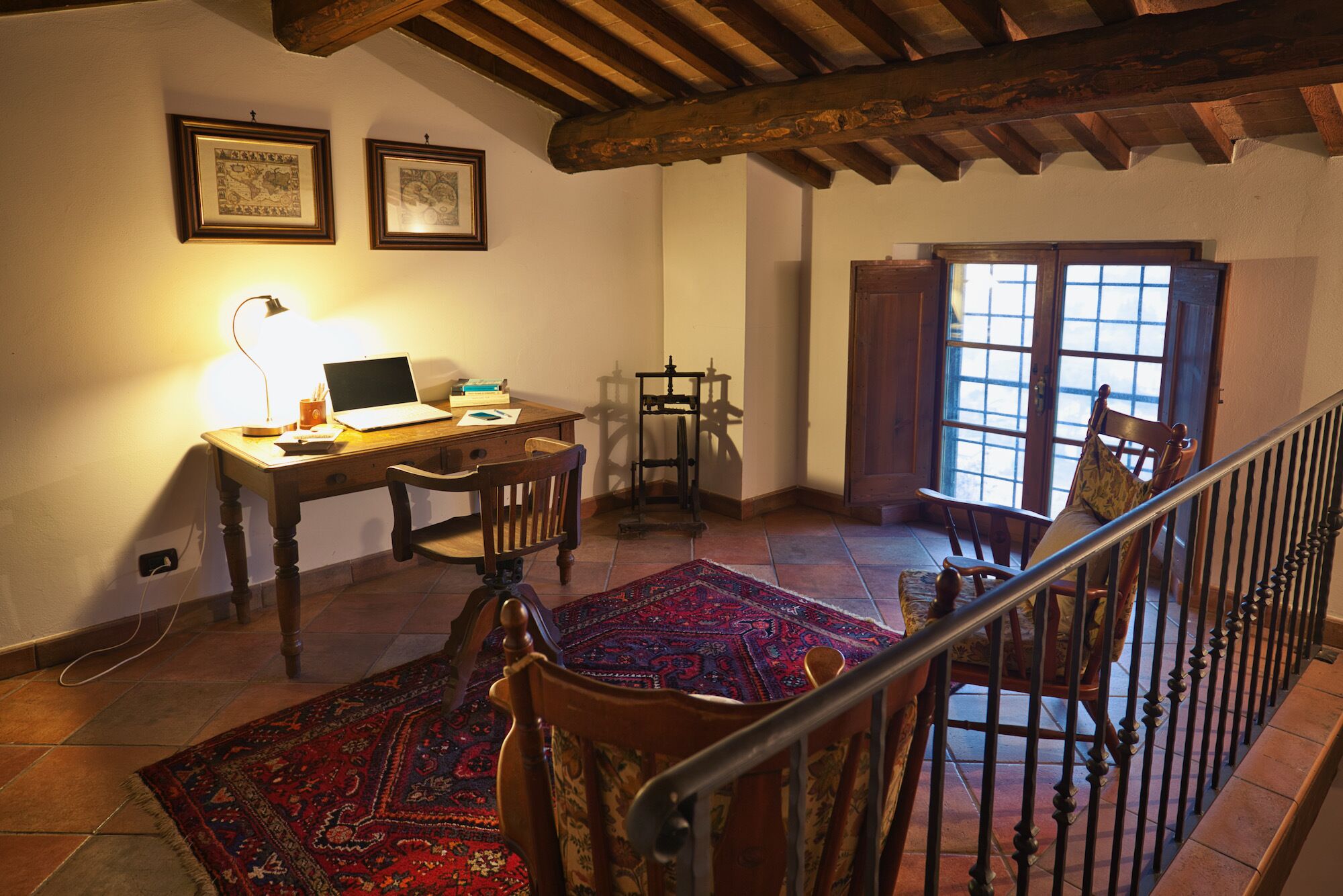 A cozy room with a wooden desk, a laptop, a chair, a patterned rug, and a staircase with a railing.