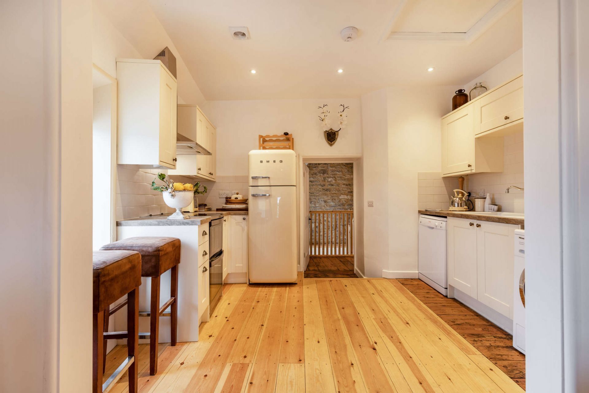 Une cuisine étroite avec un plancher en bois, des armoires blanches, un réfrigérateur et une entrée en pierre.
