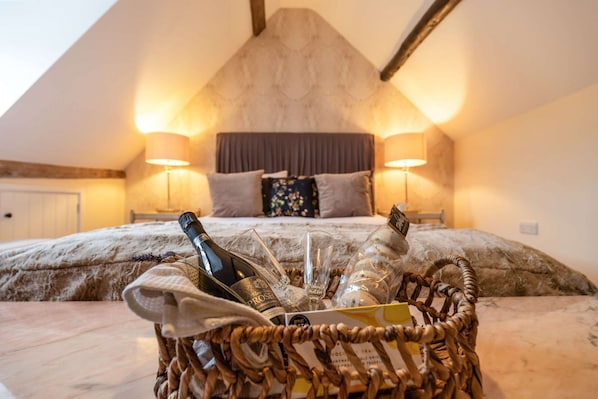 A bedroom with a bed, two bedside lamps, a wicker basket with bottles and glasses, and a headboard.