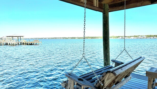 Eine Holzschaukel hängt über dem Wasser an einer Holzterrasse, im Hintergrund ein Steg und Gebäude.