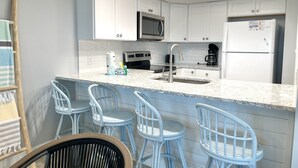 Eine moderne Küche mit weißen Schränken, einer Edelstahl-Mikrowelle, einer Kaffeemaschine und einem Kühlschrank. Es gibt drei Barhocker und einen Holzstuhl.