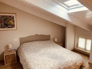 Ein Schlafzimmer mit Oberlicht, einem Bett mit gemustertem Bettbezug, einem Nachttisch mit Lampe und einem Gemälde an der Wand.