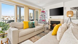 A modern living room with a beige sofa, a Christmas tree, a fireplace, and a large window overlooking a cityscape. - Stunning Water Views - Fireplace - Chelsea Harbor (Atlantic City)