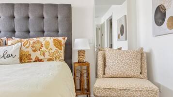 Une chambre à coucher avec une tête de lit capitonnée, un lit agrémenté d’oreillers à motifs, une table de chevet avec une lampe et une chaise avec un coussin à imprimé léopard.
