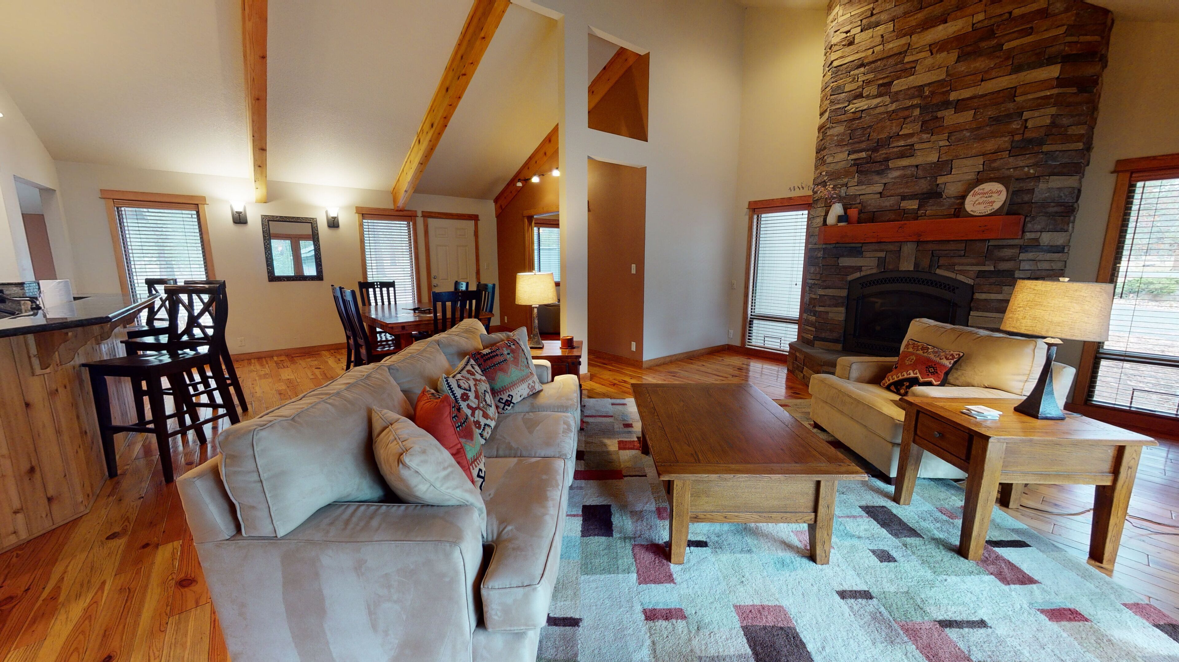 A spacious living room with a stone fireplace, wooden beams, a sofa, a coffee table, and a dining area with chairs and a table.
