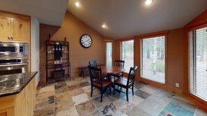 A kitchen with a built-in microwave and oven, a dining table with chairs, a clock on the wall, and large windows with blinds. - Family Oasis with Access to The Cove (Sunriver)