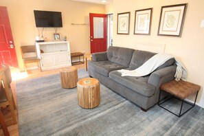 A living room with a grey sofa, two wooden stools, a red door, and a flat-screen TV mounted on the wall.