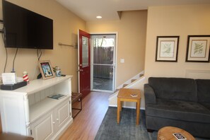 A living room with a television mounted on the wall, a red door, a grey couch, a small wooden table, and a framed picture on the wall.