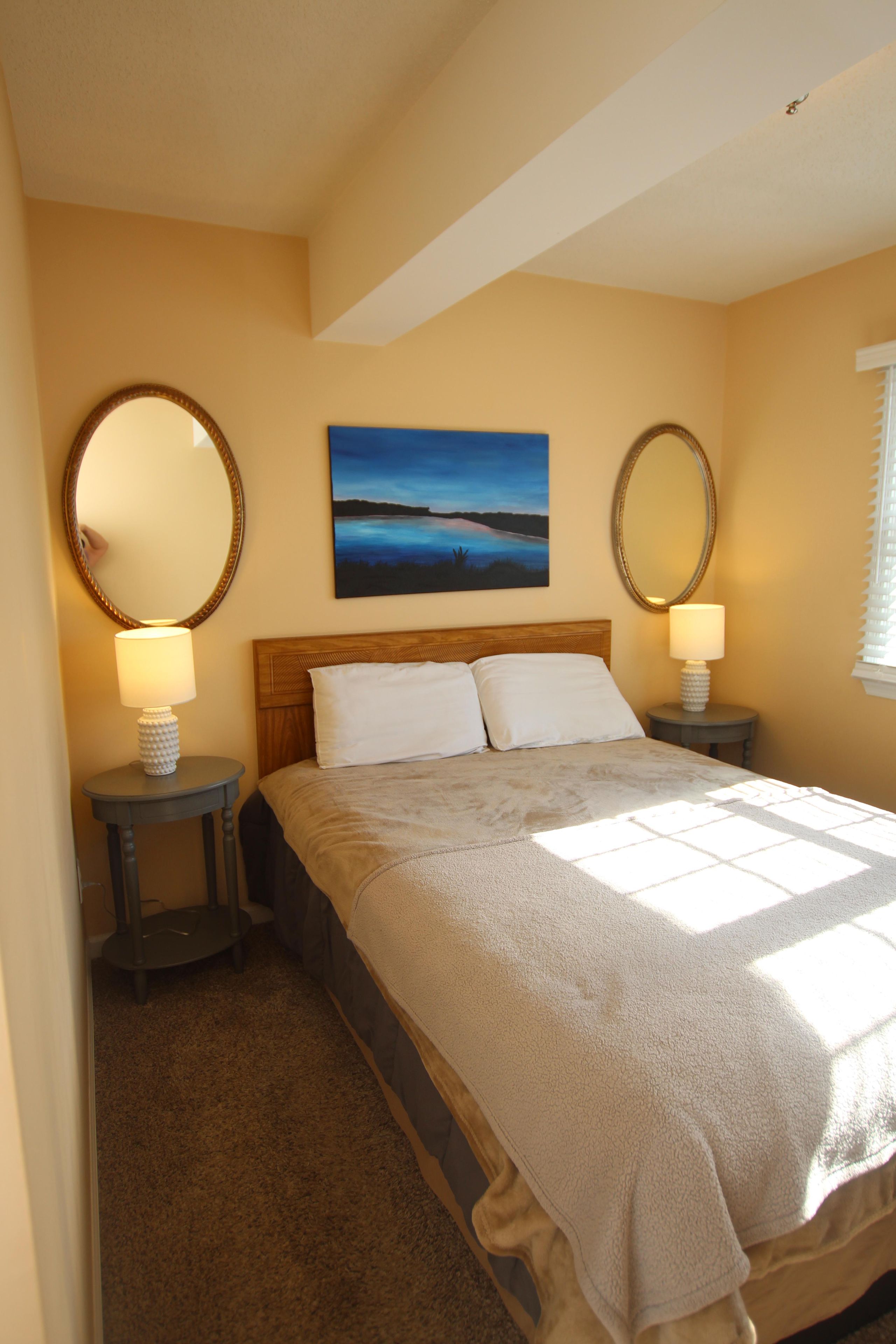 A bedroom with a large bed, two bedside tables with lamps, a painting on the wall, and a window with blinds.