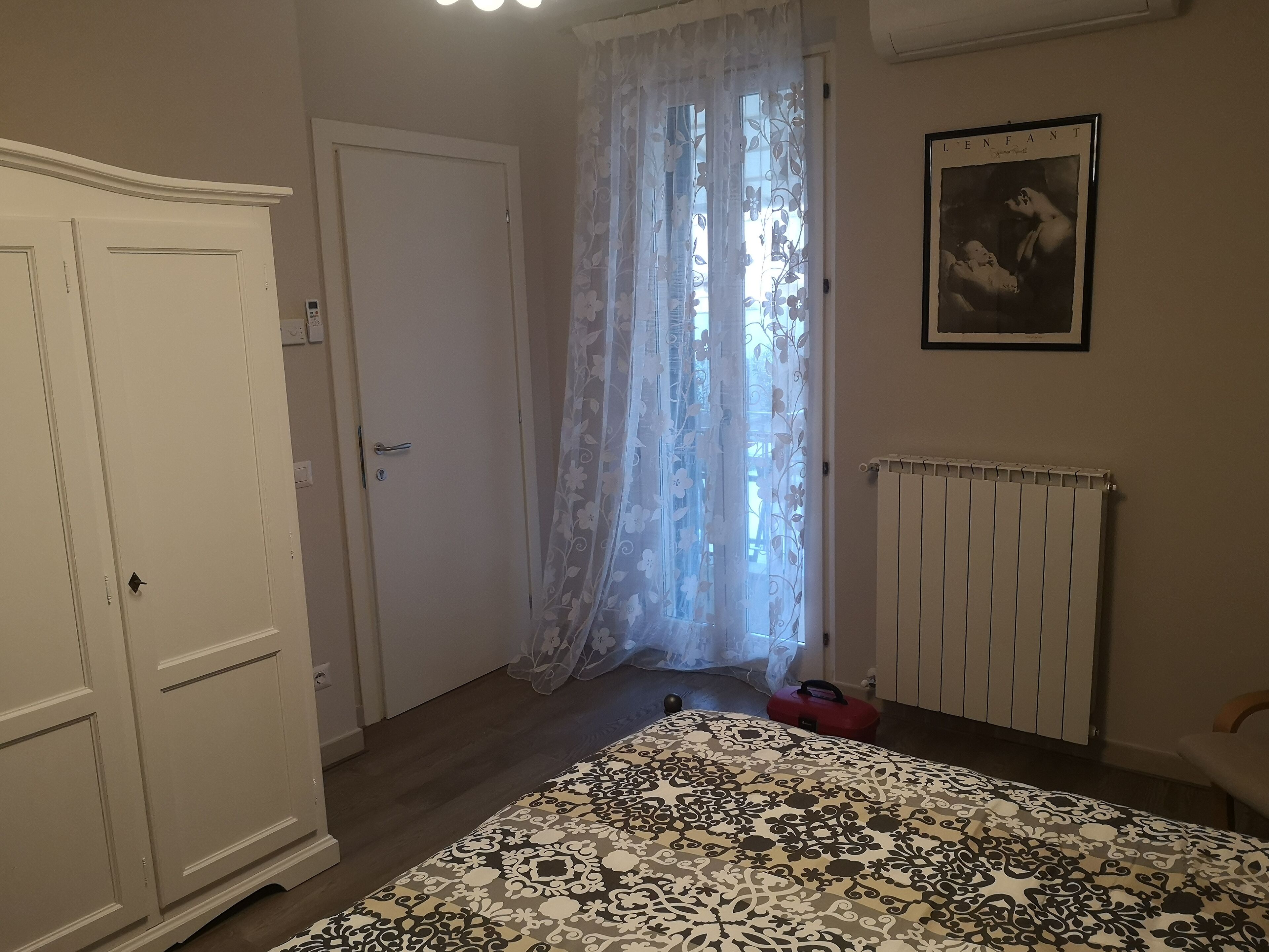 Ein Schlafzimmer mit Bett, Kleiderschrank, Heizkörper und einem Fenster mit floralem Vorhang.