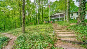 A two-story house with a porch, surrounded by a forested area with a dirt path leading to the entrance.