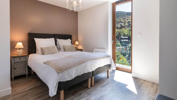 A bedroom with a large bed, two bedside tables with lamps, a chandelier, and a balcony with a view of a town and hills.