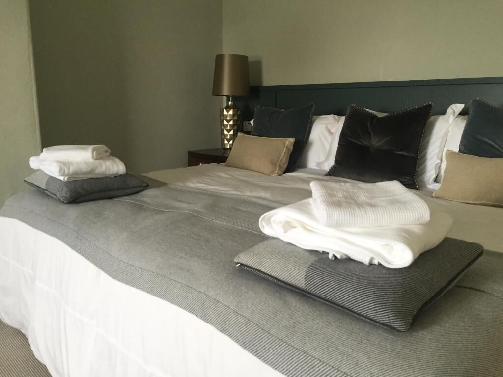 Een net opgemaakt bed met een grijs-wit dekbed, meerdere kussens en opgevouwen handdoeken op een donkergrijze kussen.