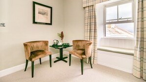 A room with two chairs, a small table, a framed picture on the wall, and a window with curtains. - Black Bull Gartmore (Stirling)