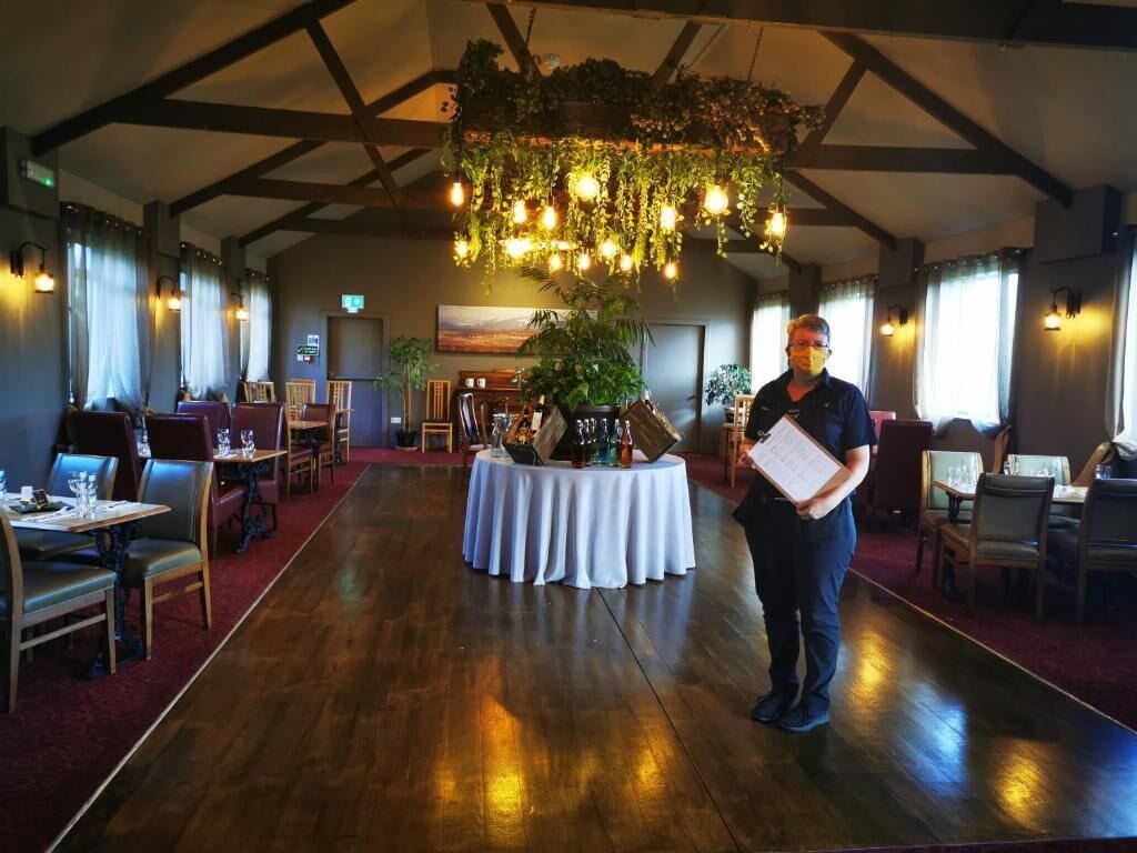 A spacious room with wooden floors, tables, and chairs arranged for dining. A large plant and hanging lights are visible in the center.