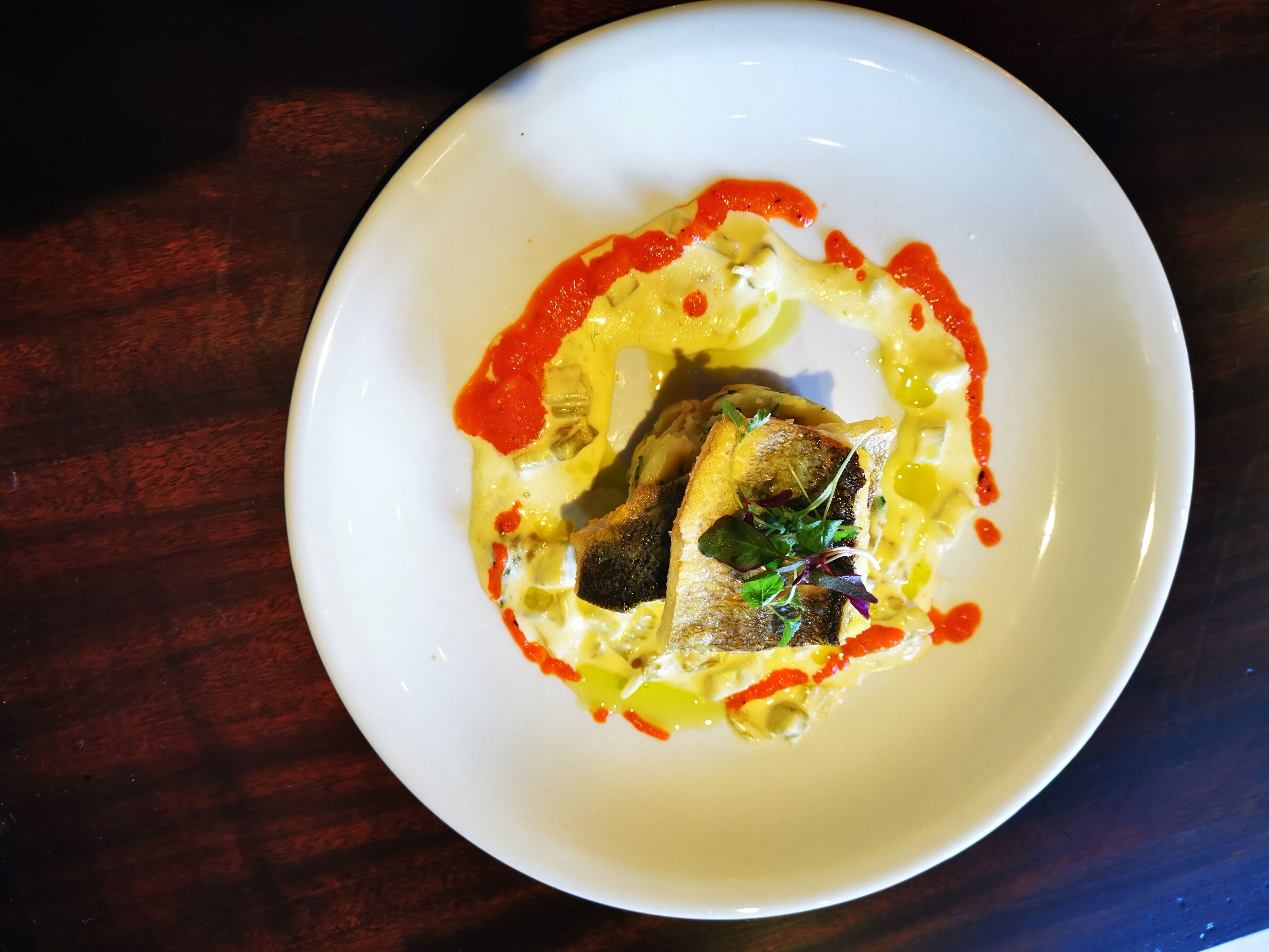 A white plate with a fish, sauce, and garnish on a dark wooden table.