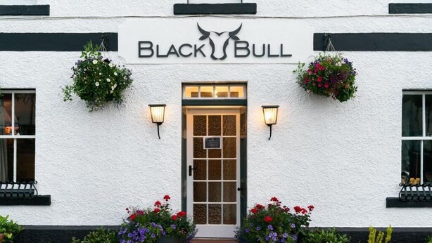 A building with a white exterior, a black and white sign reading "BLACK BULL", and a door with a glass panel. There are flower pots with red and purple flowers on either side of the entrance. - Black Bull Gartmore (Stirling)