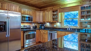 A modern kitchen with wooden cabinets, stainless steel appliances, granite countertops, and a large window with a view of trees. - Closest cabin to Tweetsie. BEST Blowing Rock Location!  Hot tub & game room (Blowing Rock)