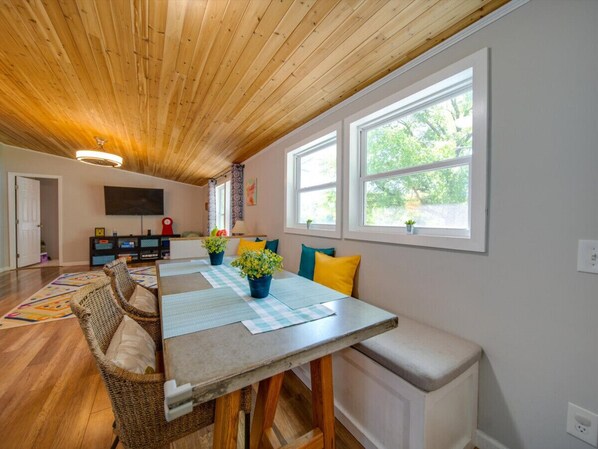 A dining area with a wooden ceiling, a long table with chairs, and a bench. There are potted plants on the table, and the room has large windows with a view of greenery outside.