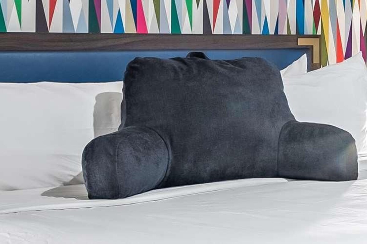Ein Bett mit weißer Bettwäsche, einem dunkelgrauen Kopfkissen und einer Wand im Hintergrund mit farbigem geometrischen Muster.