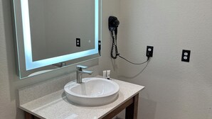 Een badkamer met een witte wastafel, een houten opbergmeubel, een grote spiegel met LED-verlichting en een haardroger aan de muur.