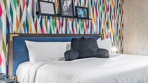 Een hotelkamer met een bed, wit beddengoed, een blauw hoofdeinde en een wand met een kleurrijk geometrisch patroon.
