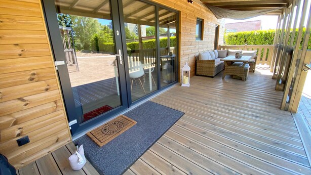 A wooden deck with a covered seating area, a welcome mat, and a glass door leading outside. - Wassermann vacation home with box-spring beds and large terrace (Wunstorf)